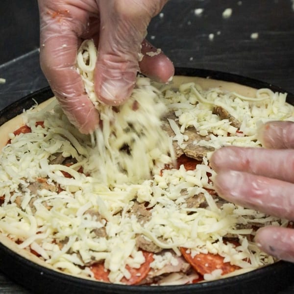 Gloved hands sprinkle shredded cheese over a deep-dish pizza with pepperoni and meat toppings.