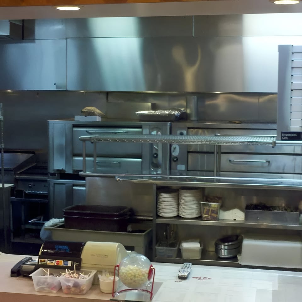 A commercial kitchen with stainless steel appliances, shelves of plates, and a counter with a cash register.